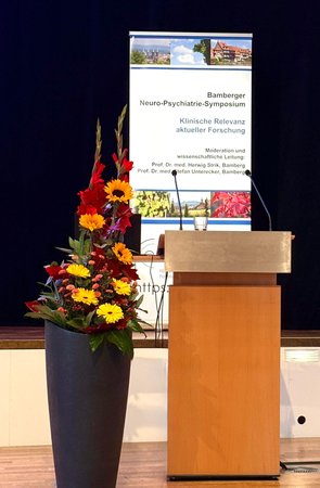 Rednerpult mit Blumen und im Hintergrund das RollUp vom Bamberger Neuro-Psychiatrie-Symposium